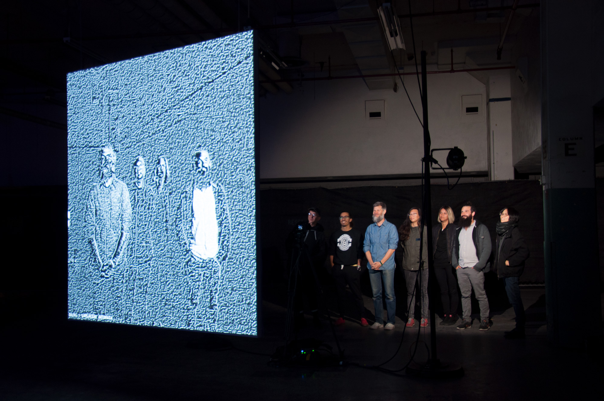 Seven people looking at a filtered projection of themselves on a screen.