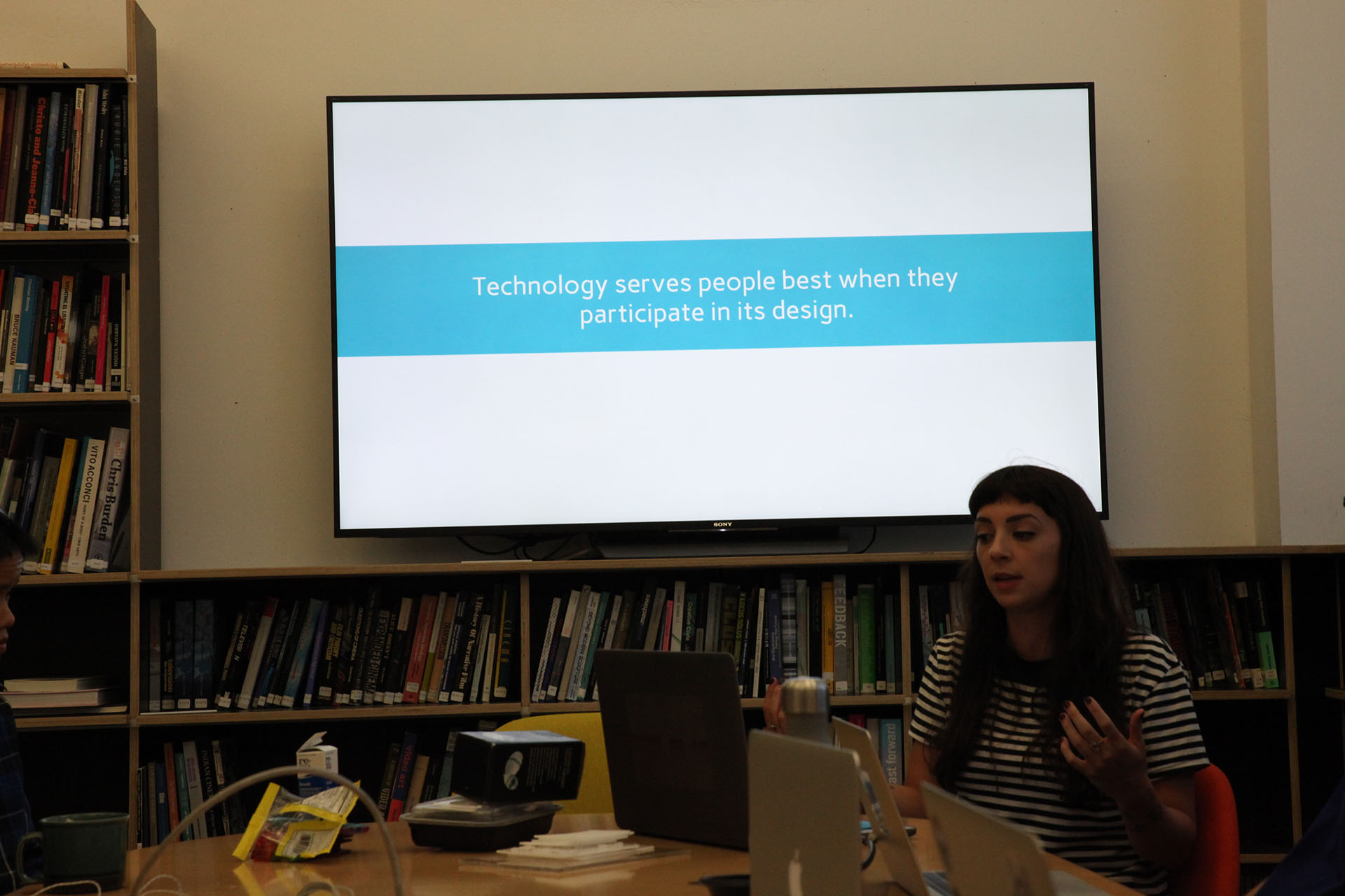 A woman giving a presentation in front of a TV. The presentation on the screen reads 'Technology serves people best when they participate in its design.'