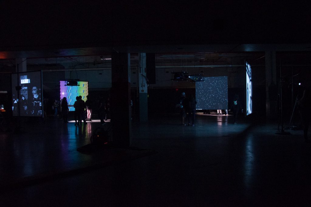 A large dark room. People are standing in front of large screens with interesting projections.